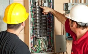 Two electricians in hard hats inspecting a residential electrical panel with exposed circuit breakers and wiring.